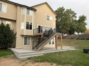A two-story tan house with a new wooden deck and stairs leading down to a concrete patio in a backyard.