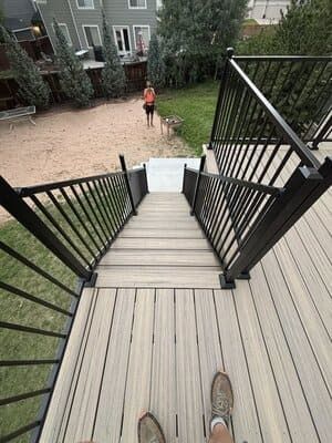 A first-person view looking down a wooden deck staircase leading to a backyard where a person stands on a sandy patch.