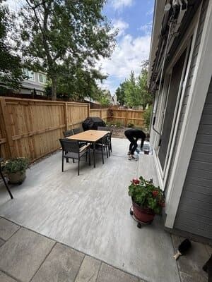 A person paints a gray patio near a wooden fence and house, with a dining table and potted plants on the terrace.