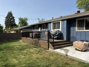 A blue house with a dark brown deck, black railings, and a concrete patio in a backyard with a wooden fence.