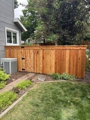 A new, tall, vertical-plank wood fence with a gate stands in a residential backyard next to a grey house with an AC unit.