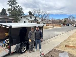 Two individuals in work attire stand next to an open black cargo trailer on a residential street.