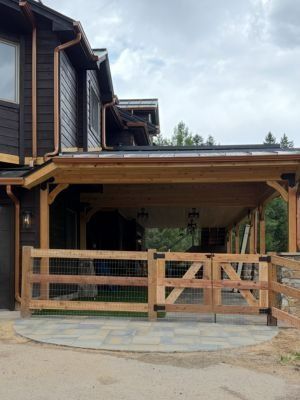 A wooden farm-style gate and fence line a stone patio under a timber-framed porch attached to a dark-sided house.