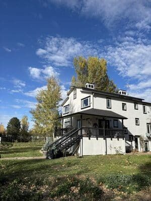 A three-story white house with a dark roof and porch stairs, set against a blue sky with autumn trees.