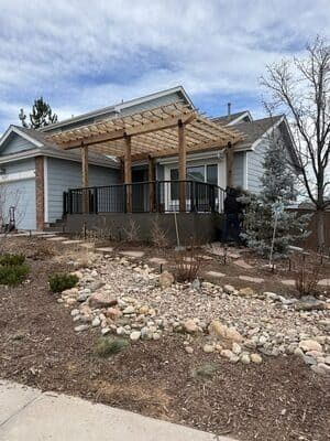 A house with a large wooden pergola over a deck, featuring black railings, a stone facade, and a rocky landscaping bed.