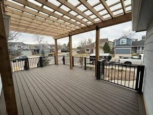 A wood-plank deck with a pergola overhead, featuring black metal railings and a view of a suburban neighborhood.