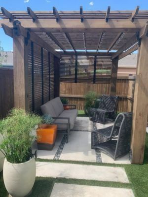 A wooden pergola covers an outdoor seating area with a sofa, two chairs, and an orange accent table on a paved patio.