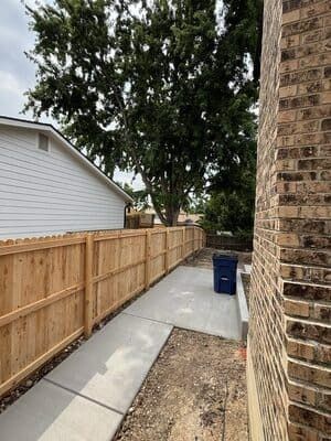 A narrow concrete walkway between a wooden fence and a brick house wall, with a blue trash bin in the background.
