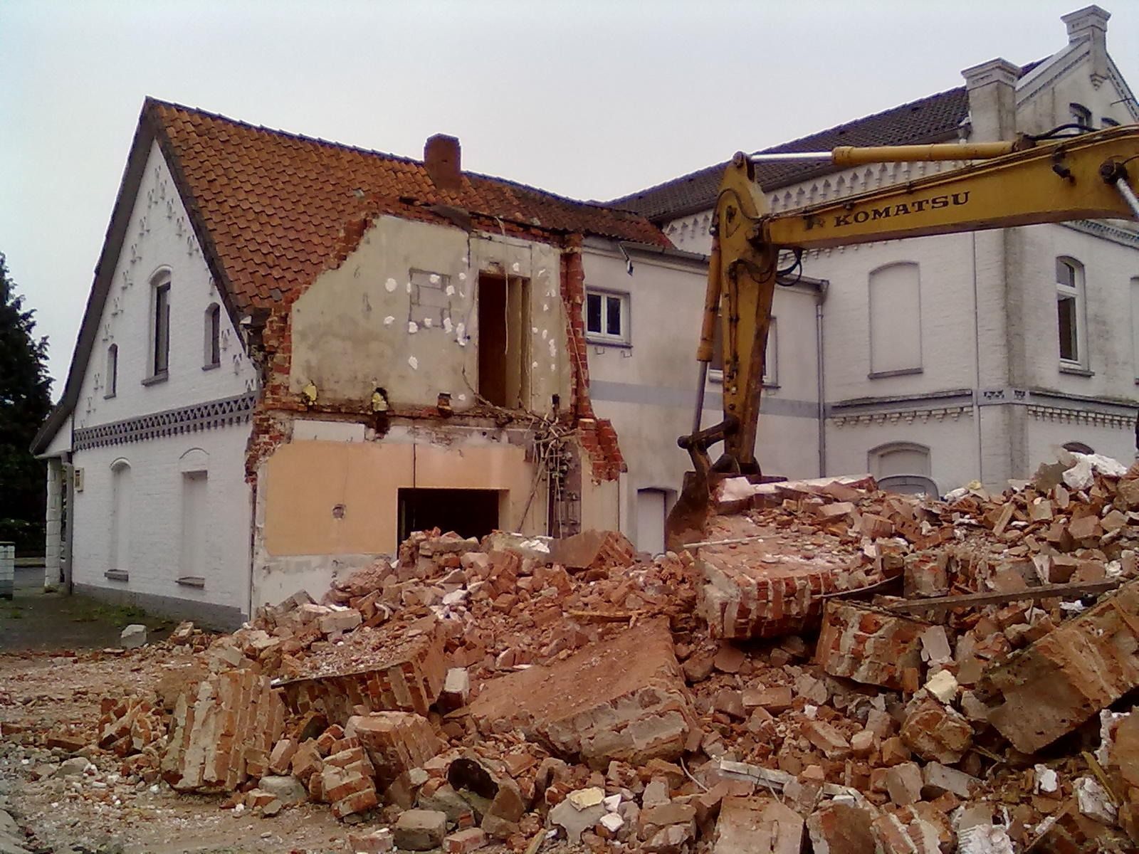 House being demolished by an excavator, with rubble piled in front of a partially destroyed building