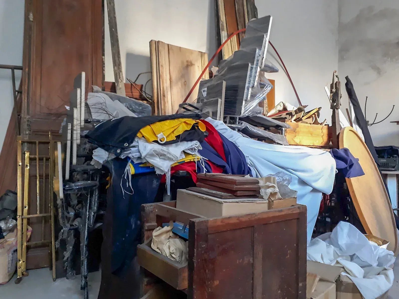 Cluttered storage area with stacked wood, metal sheets, furniture, and piles of fabric and debris