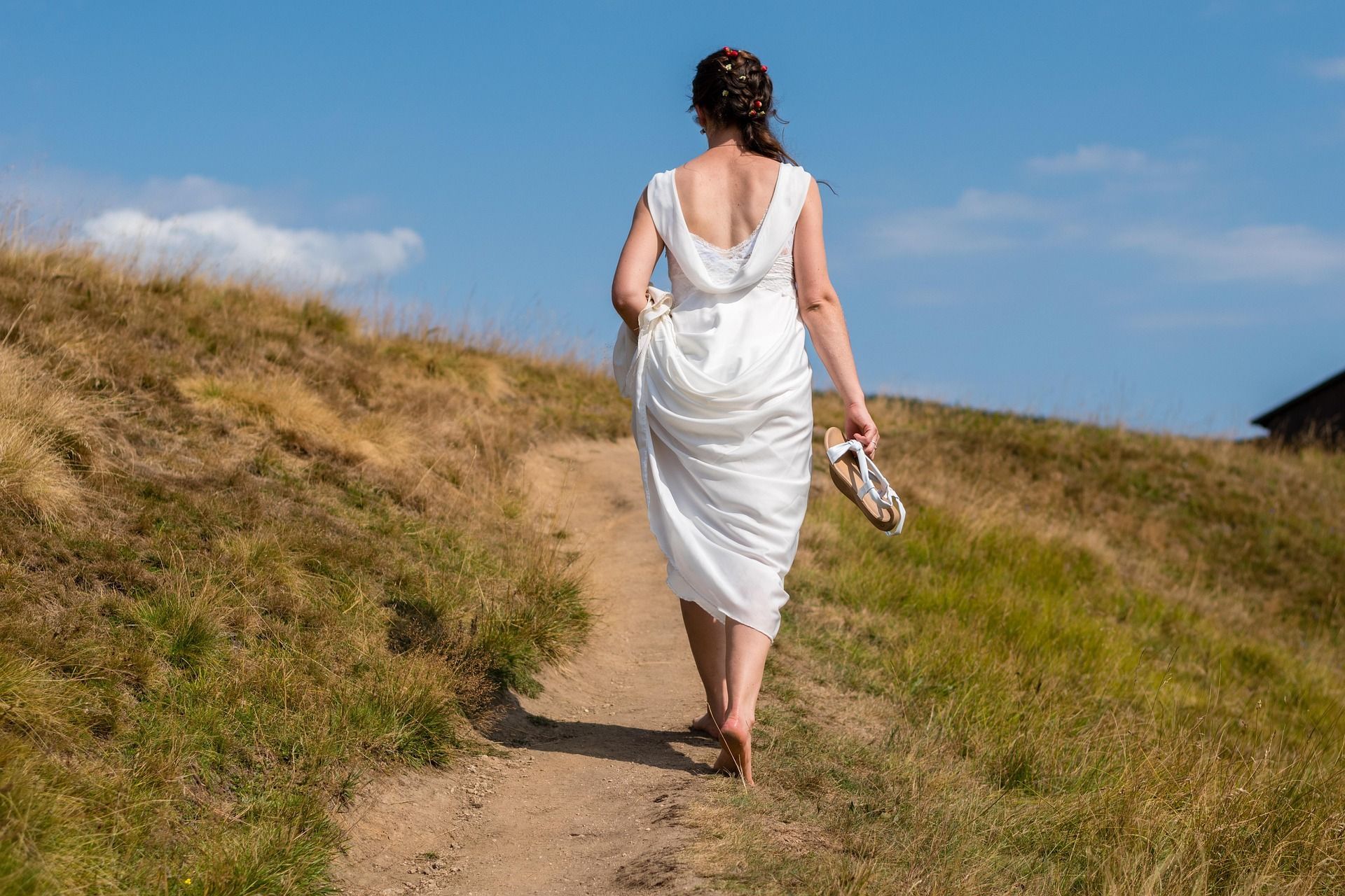 Vrouw in een witte jurk loopt over een zandpad op een grasheuvel onder een blauwe hemel.