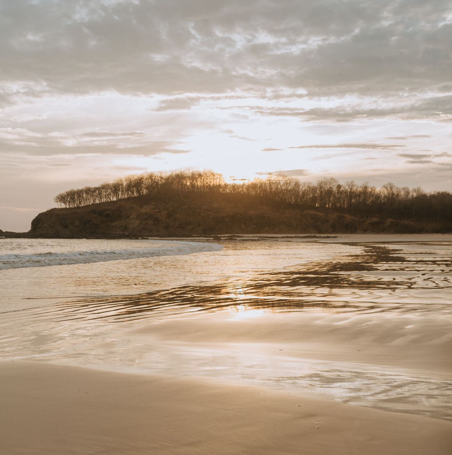 The sun is setting over a beach with a small island in the distance.
