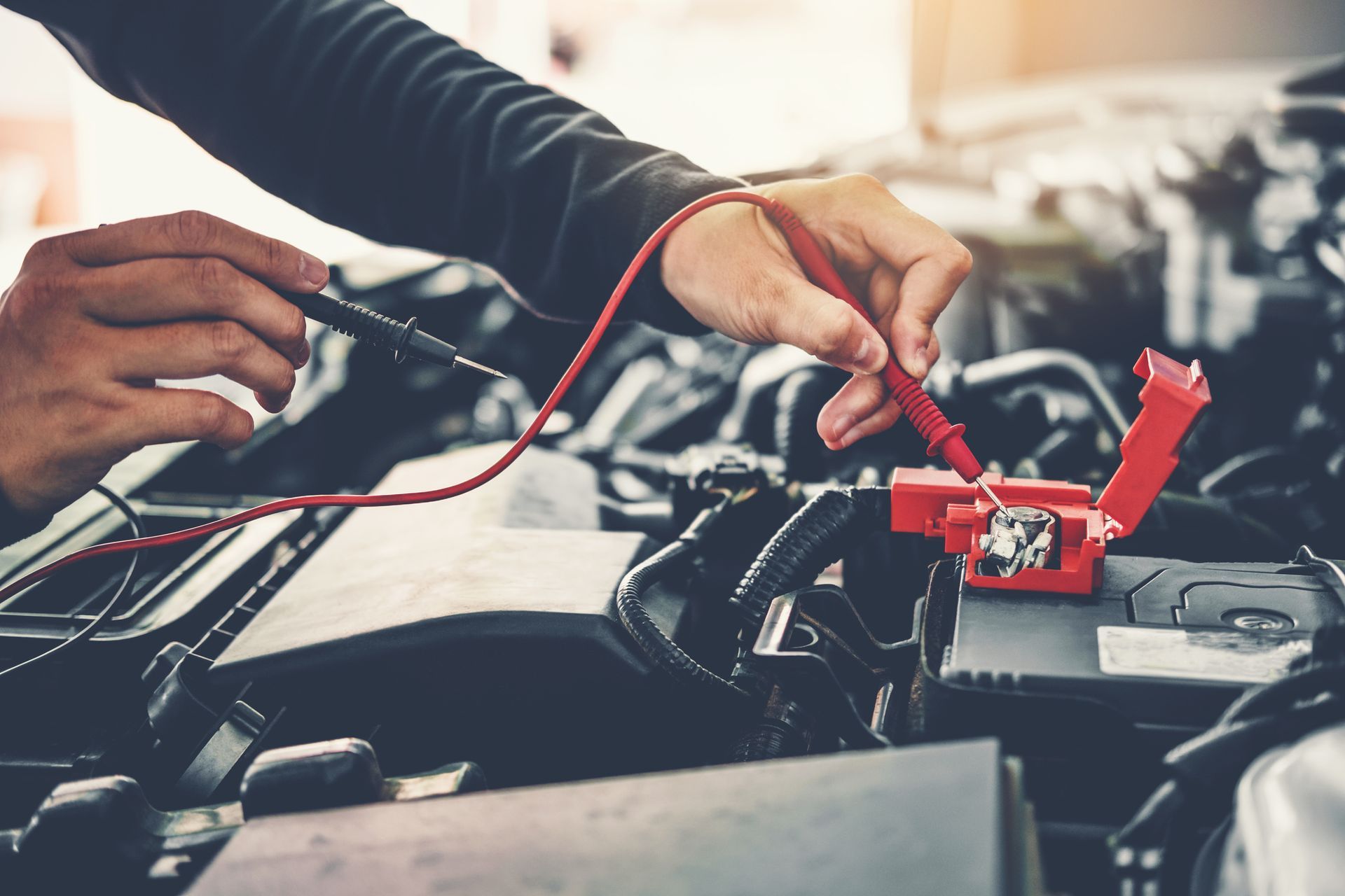 Hands using a multimeter to test a car battery in an engine bay. Red and black wires are visible.