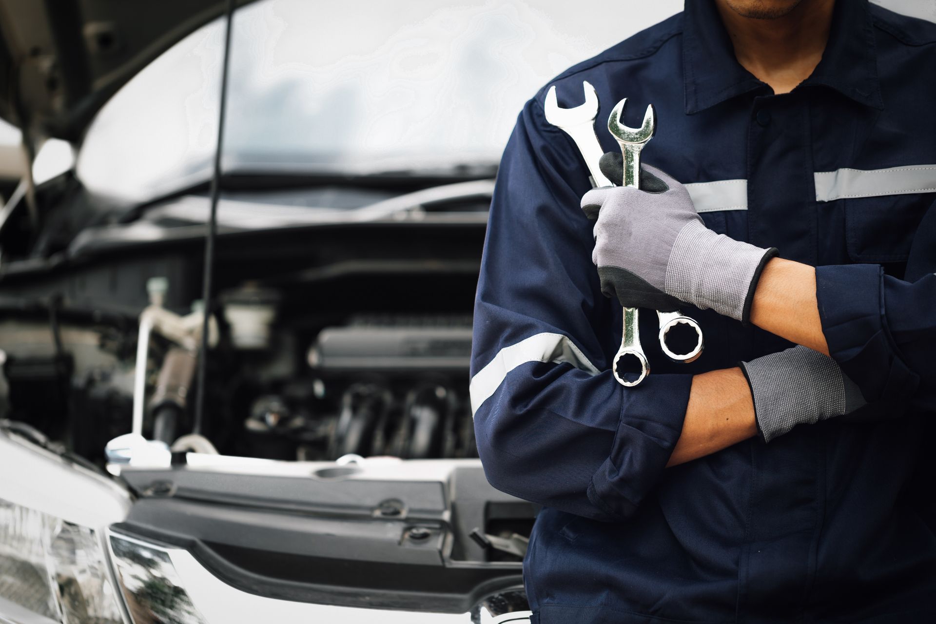 Mechanic holding wrenches, wearing blue uniform and work gloves, in front of a car with an open hood.