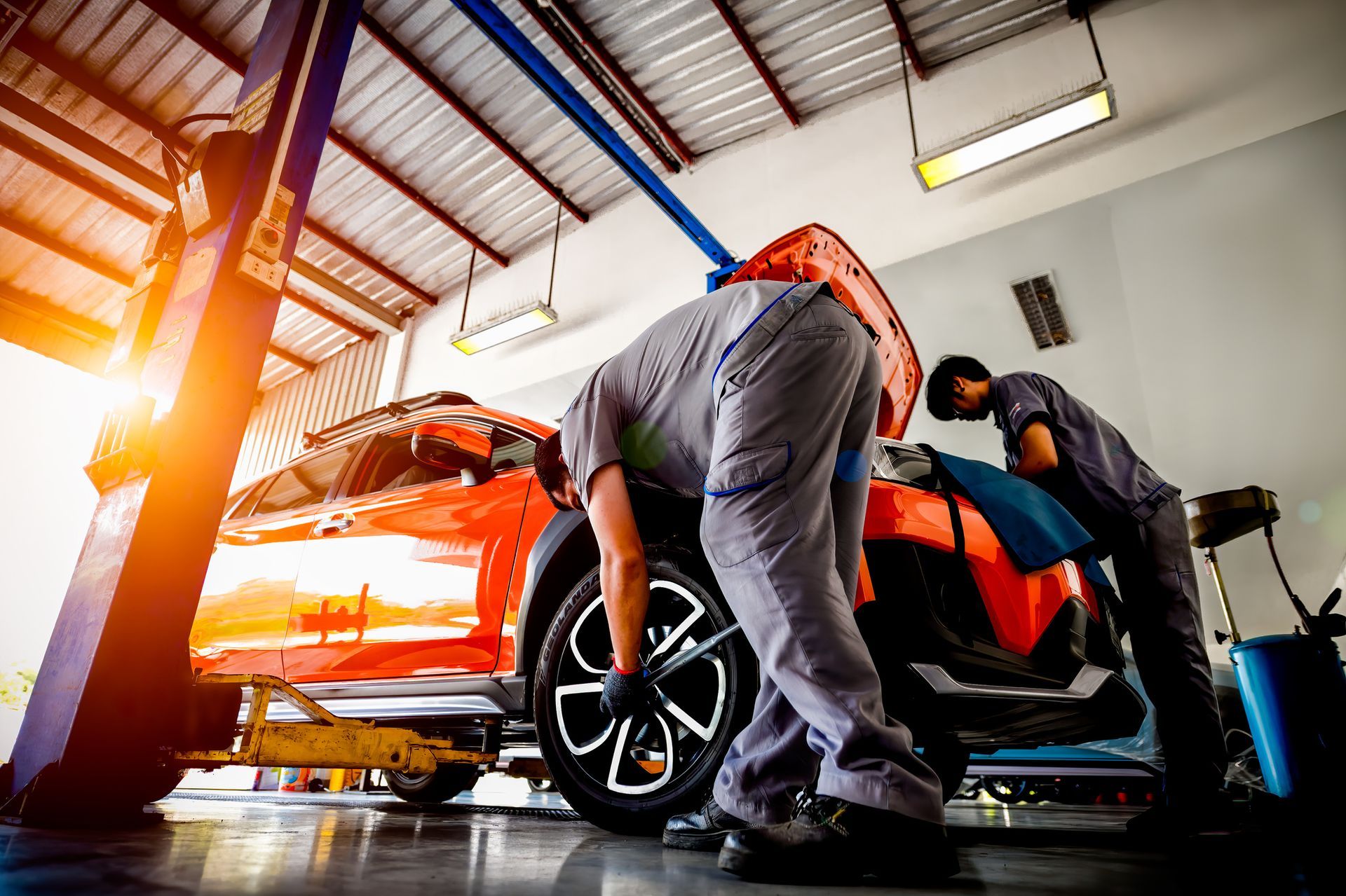Mechanics working on a red car in a well-lit auto shop, one removing a wheel, the other under the hood.