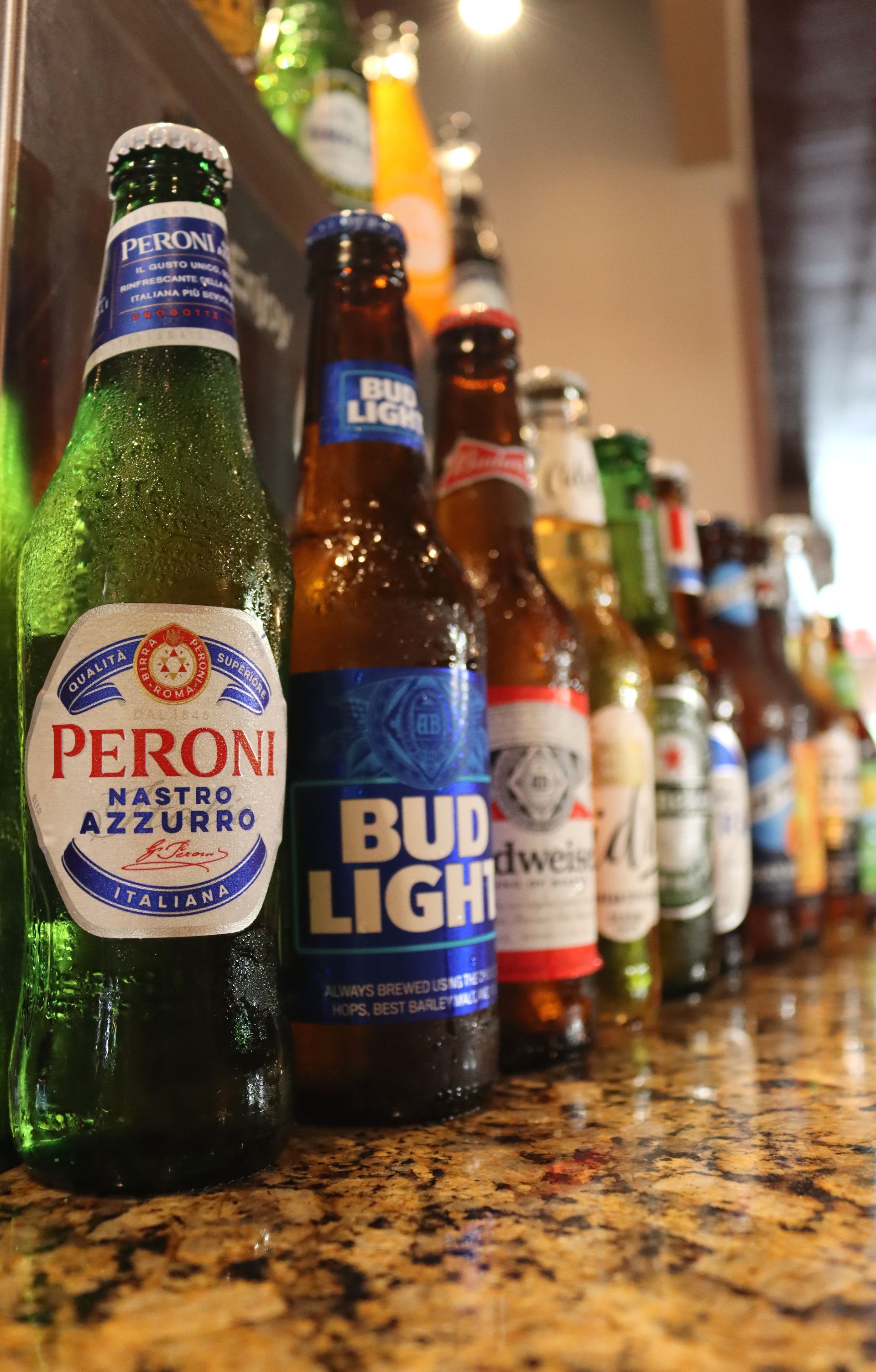 A row of beer bottles including peroni and bud light are lined up on a counter.