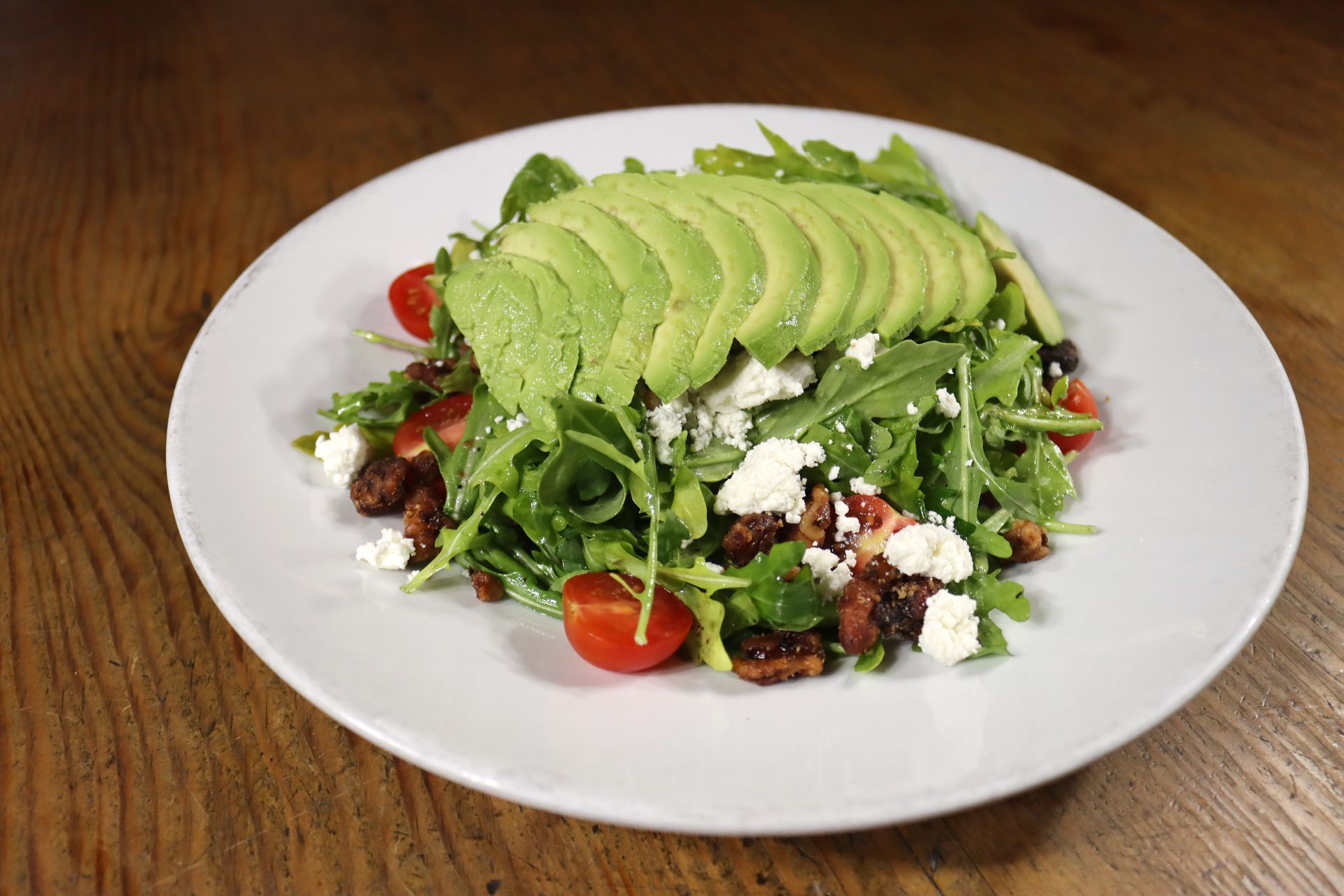 A salad with avocado , tomatoes , and feta cheese on a white plate on a wooden table.