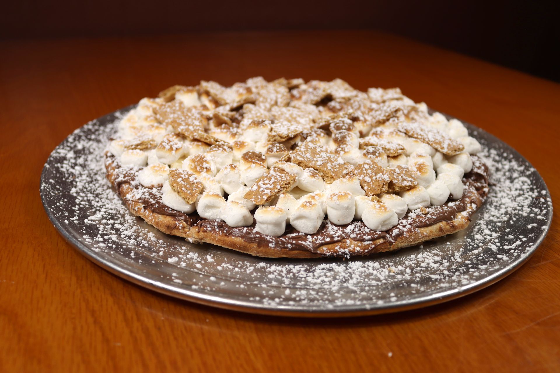 A s'mores pizza with marshmallows and graham crackers on a pizza pan on a wooden table.