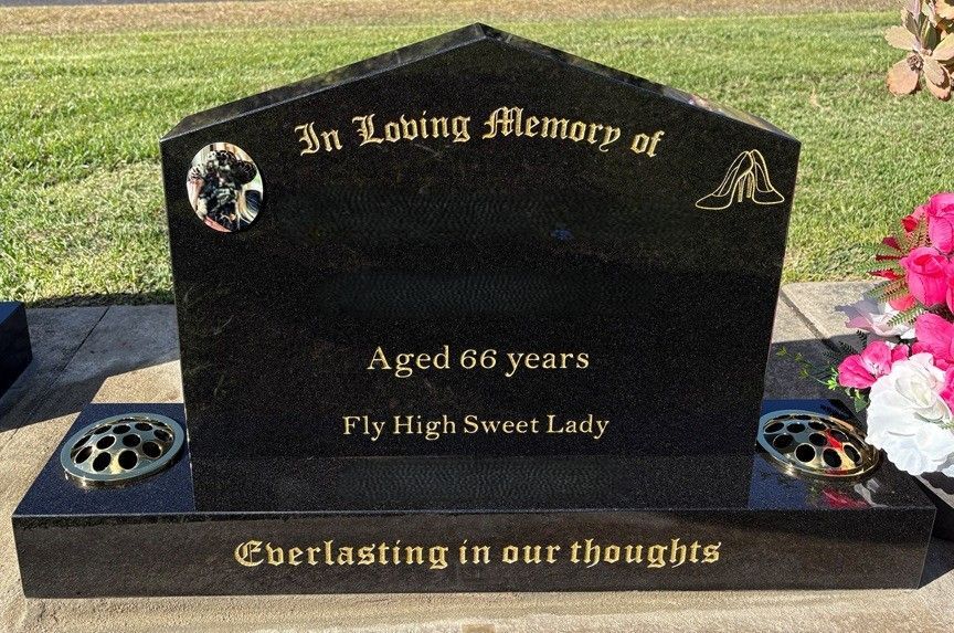 Black headstone with golden text; reads "In Loving Memory," aged 66, "Fly High Sweet Lady," and "Everlasting in our thoughts."