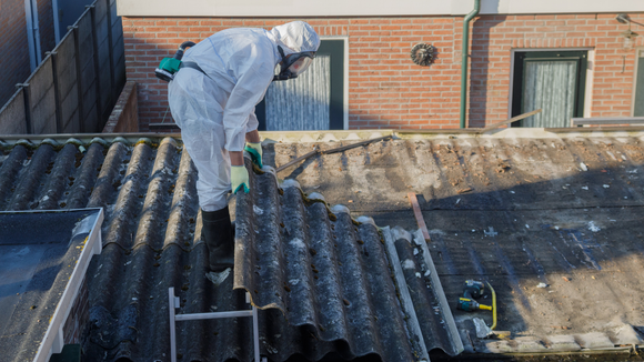 A person in a protective suit and respirator removes corrugated roofing sheets from a residential building.