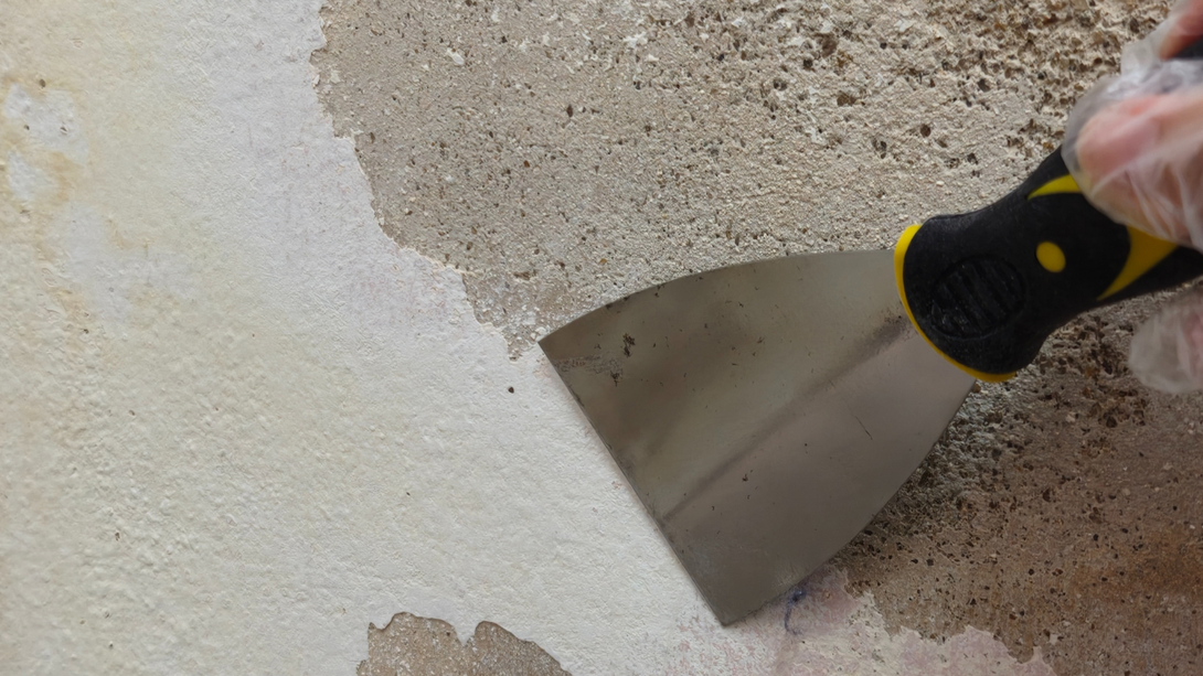 A gloved hand holds a metal putty knife scraping loose plaster off a rough concrete wall.