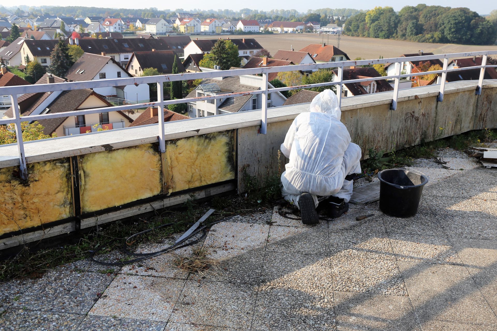 A person in white protective gear kneels on a rooftop, working near a yellow-paneled railing overlooking a suburban town.