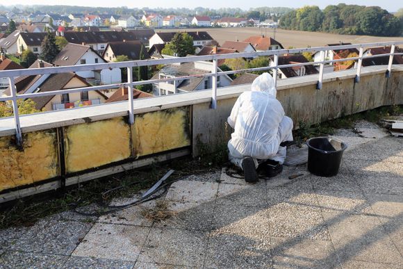 A person in white protective gear kneels on a rooftop, working on the side of a balcony overlooking a residential area.