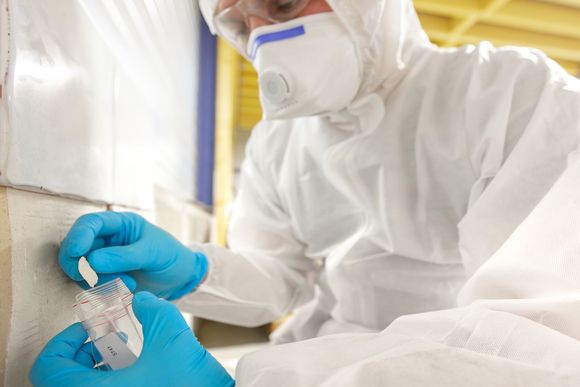 A person in a full white hazmat suit and respirator uses blue gloves to place a sample into a small collection container.