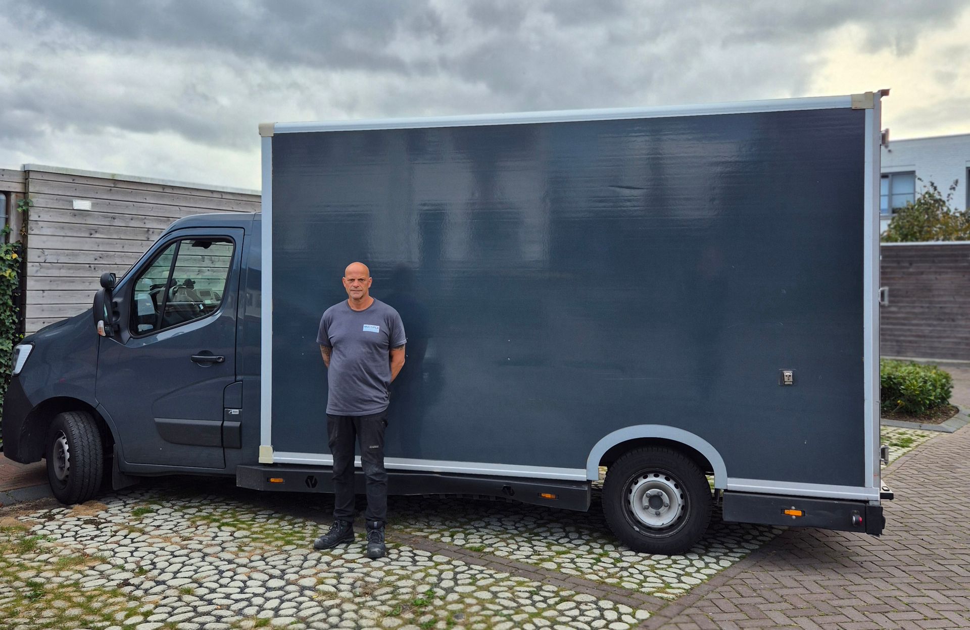 Een man staat naast een donkergrijze bestelbus; bewolkte dag, buiten.