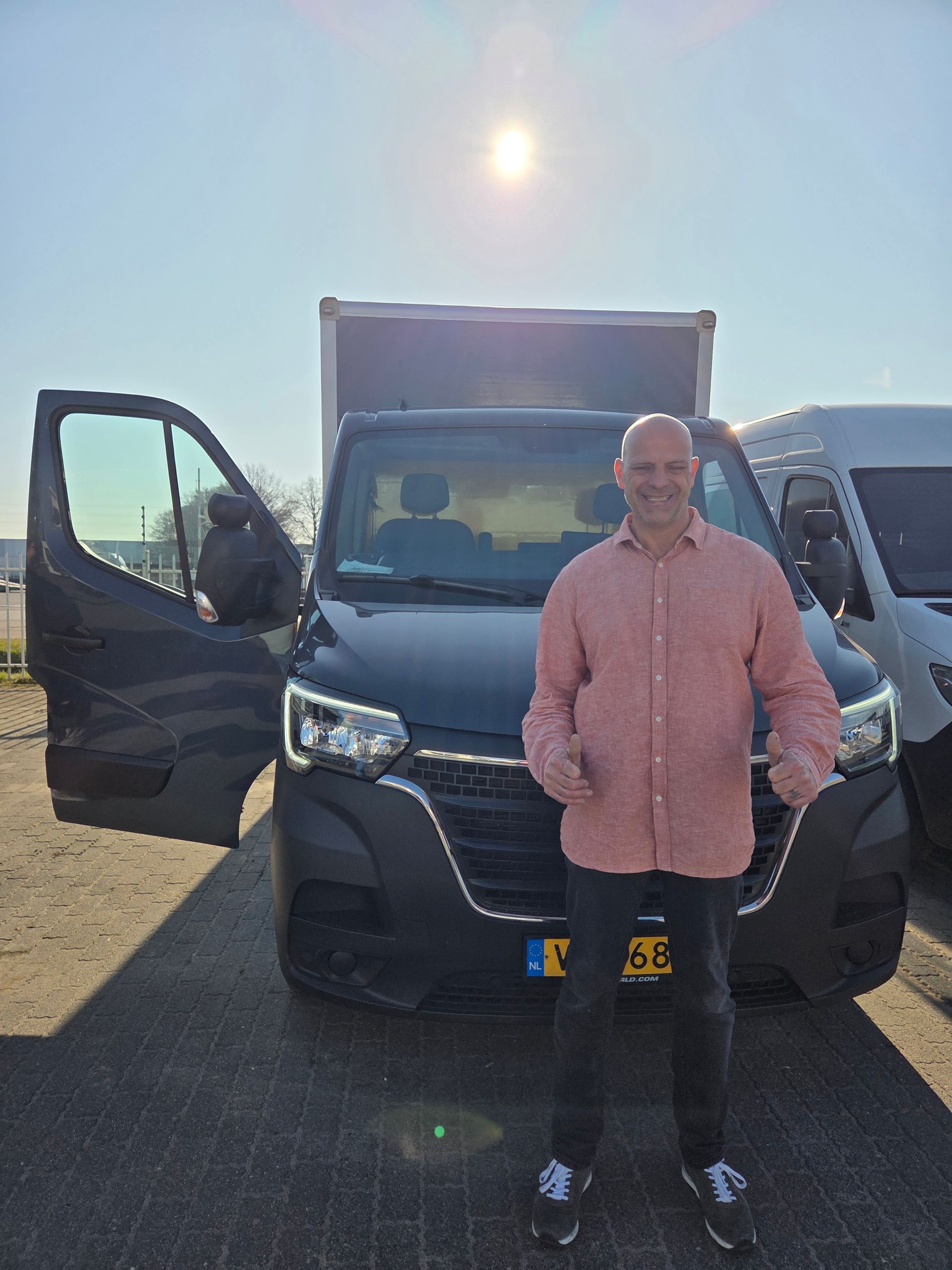 Een man staat naast een zwarte bestelbus met een open deur op een zonnige parkeerplaats.