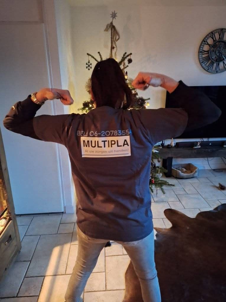 Een persoon die zijn armen spant, een shirt draagt met "Multipla" op de rug, en voor een kleine kerstboom en een klok staat.