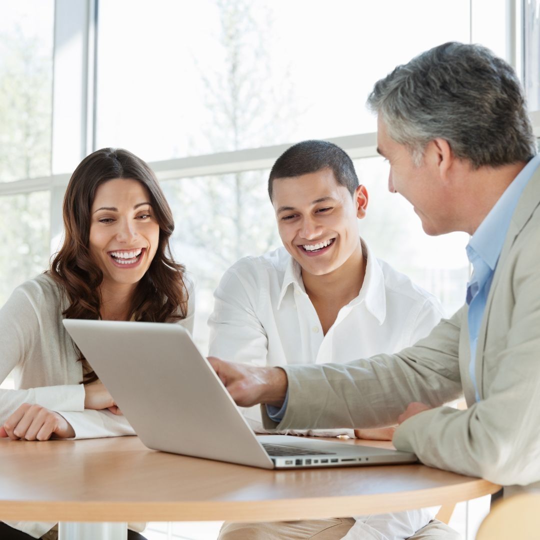 Three people smile while looking at a laptop at a table in a bright room.