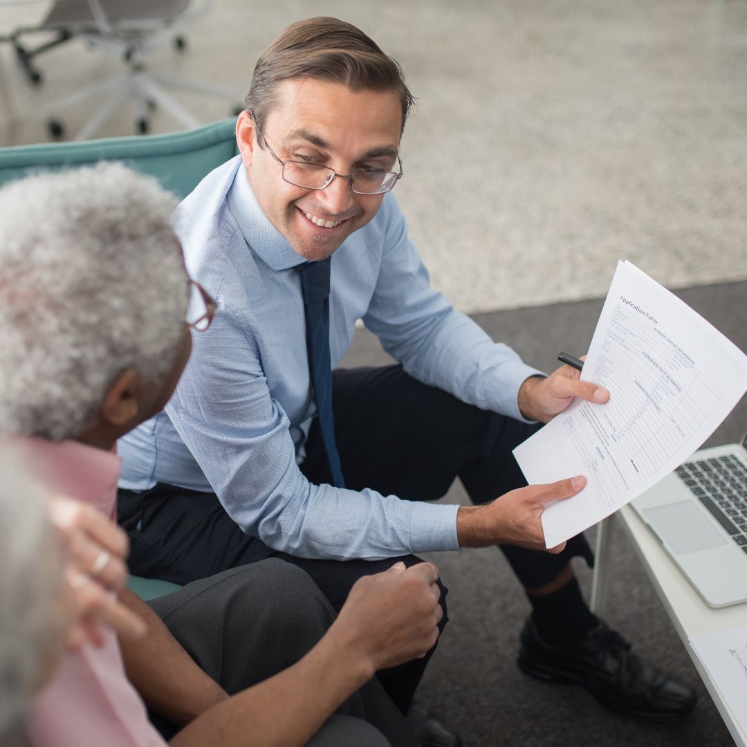 Man in suit reviews documents with seated person. Laptop on table.