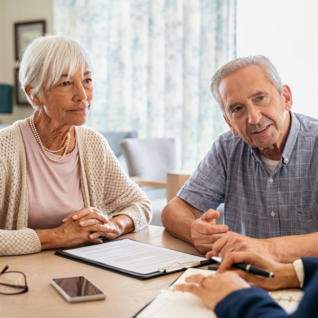 Senior couple at a table, looking at documents. A hand holding a pen points to a paper.
