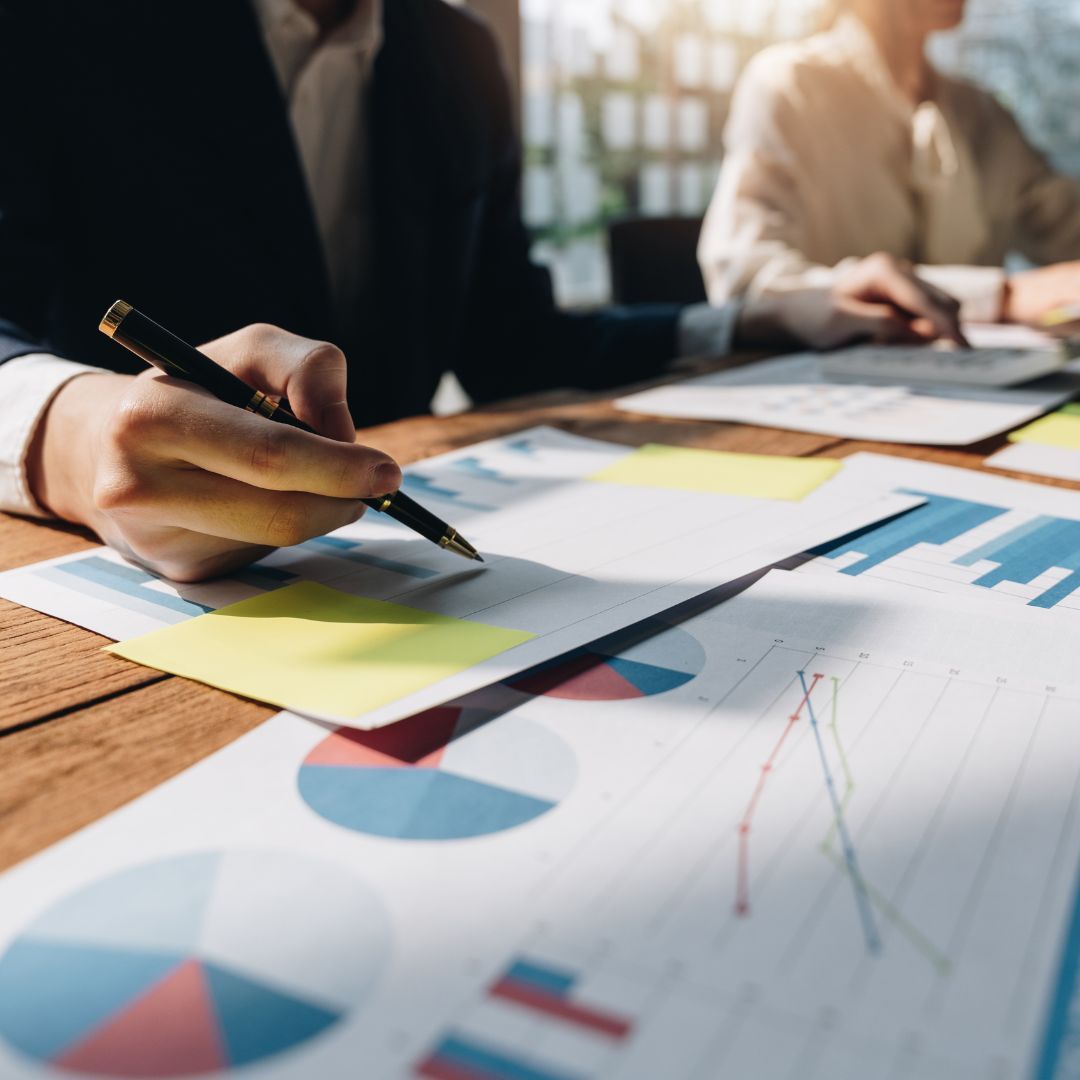 Person in suit writing on a chart with graphs at a table with another person, yellow sticky notes.
