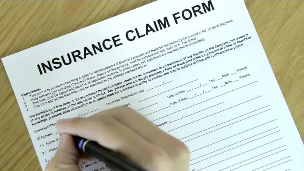 Person filling out an insurance claim form with a blue pen on a wooden desk.