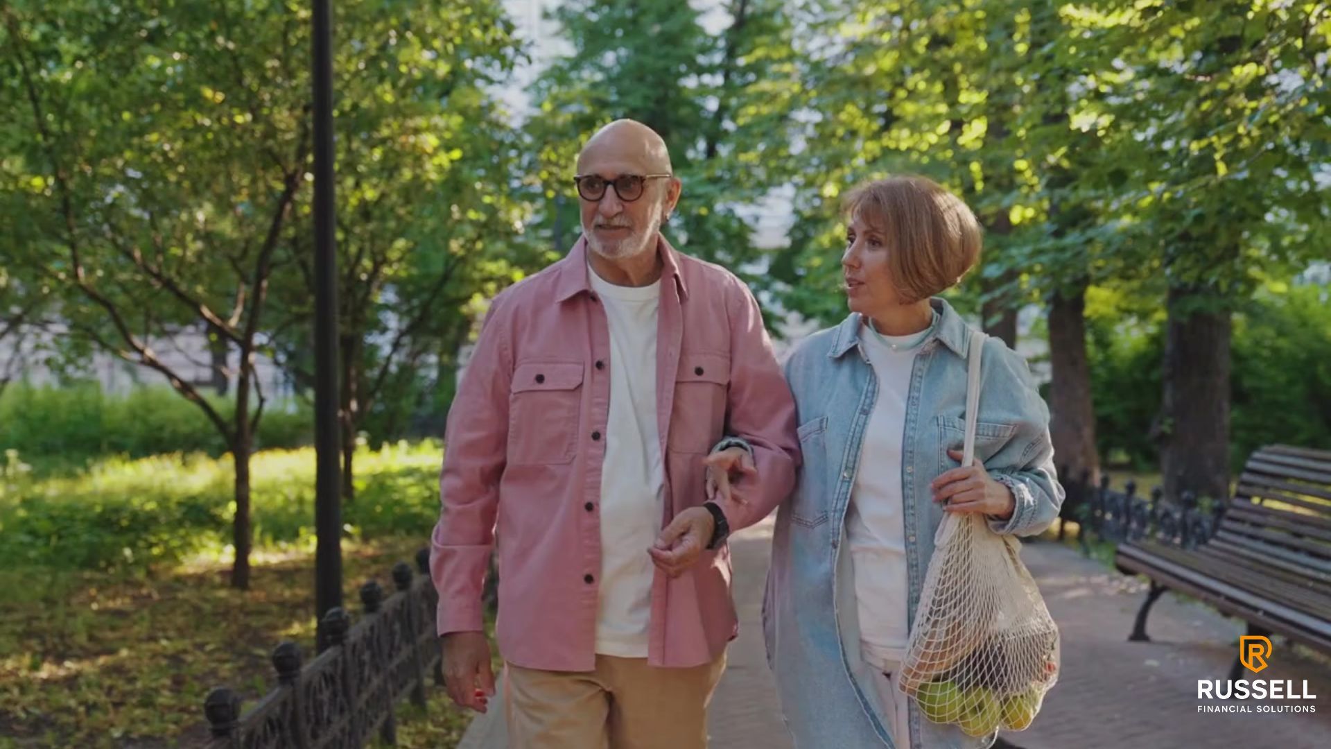An older couple walks arm-in-arm in a park, smiling. The man wears pink, the woman light blue.