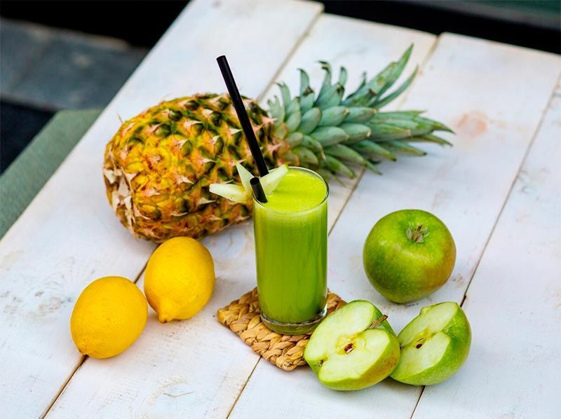 A pineapple , apples , lemons and a glass of green juice on a table.