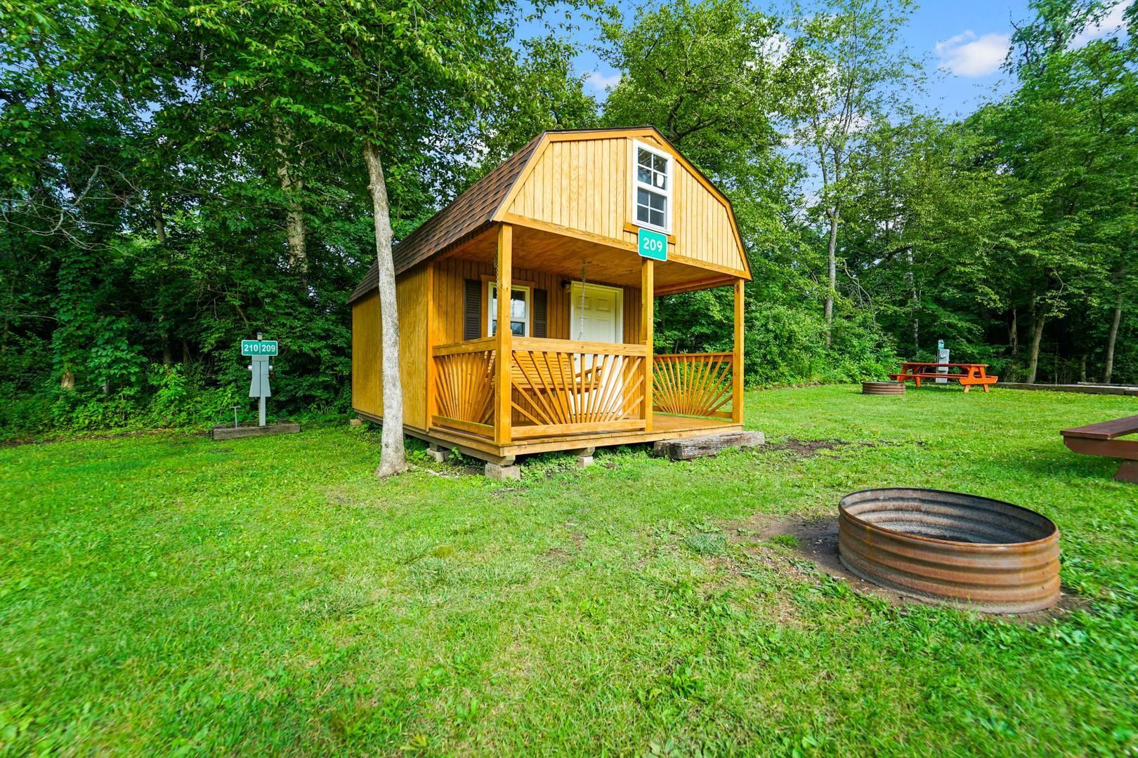 A small wooden cabin with a front porch and a metal fire ring sits in a grassy, wooded campsite under a clear blue sky.