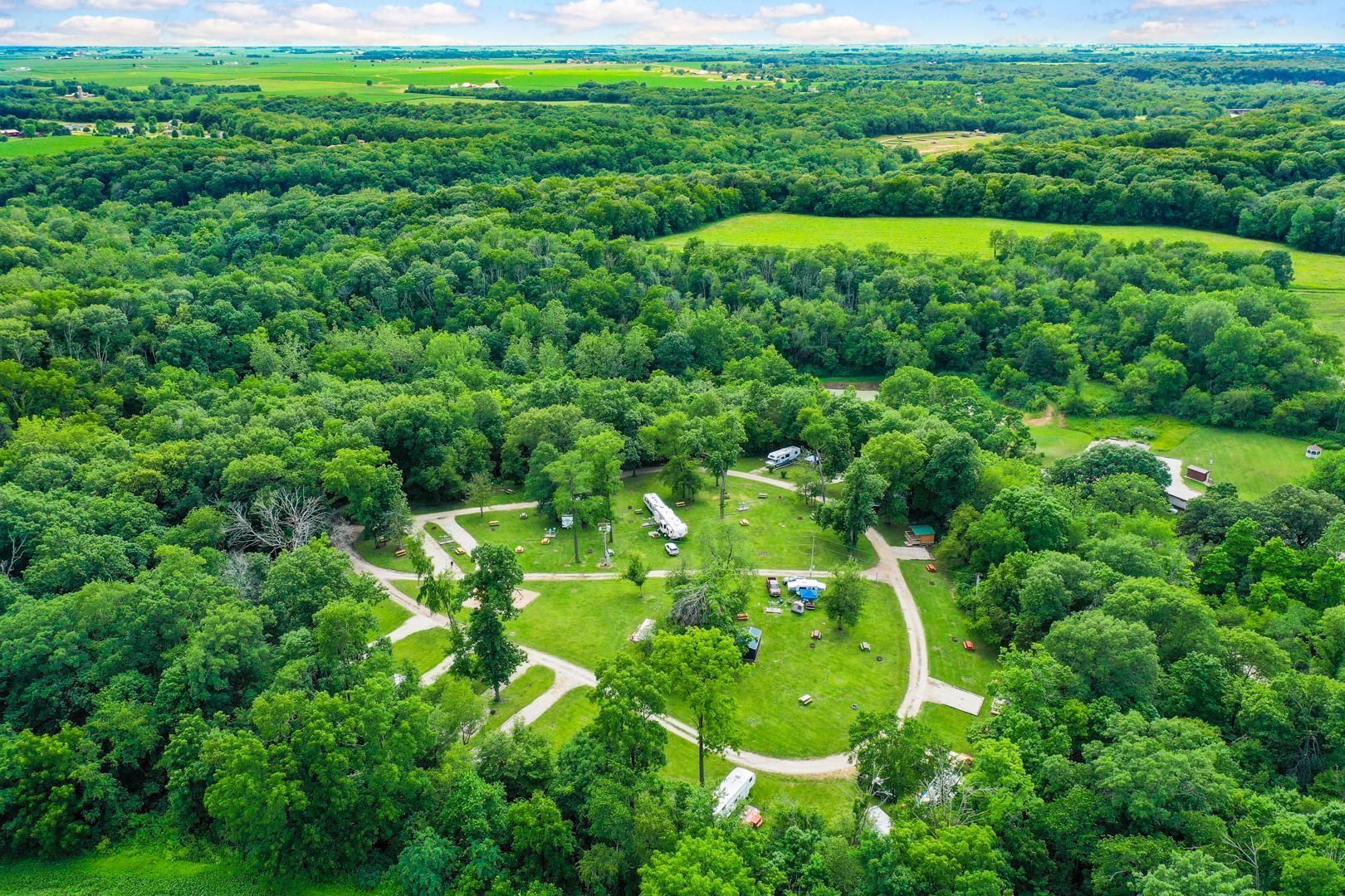 Aerial view of a small campground nestled within a lush forest, featuring gravel roads and scattered RVs on grassy sites.