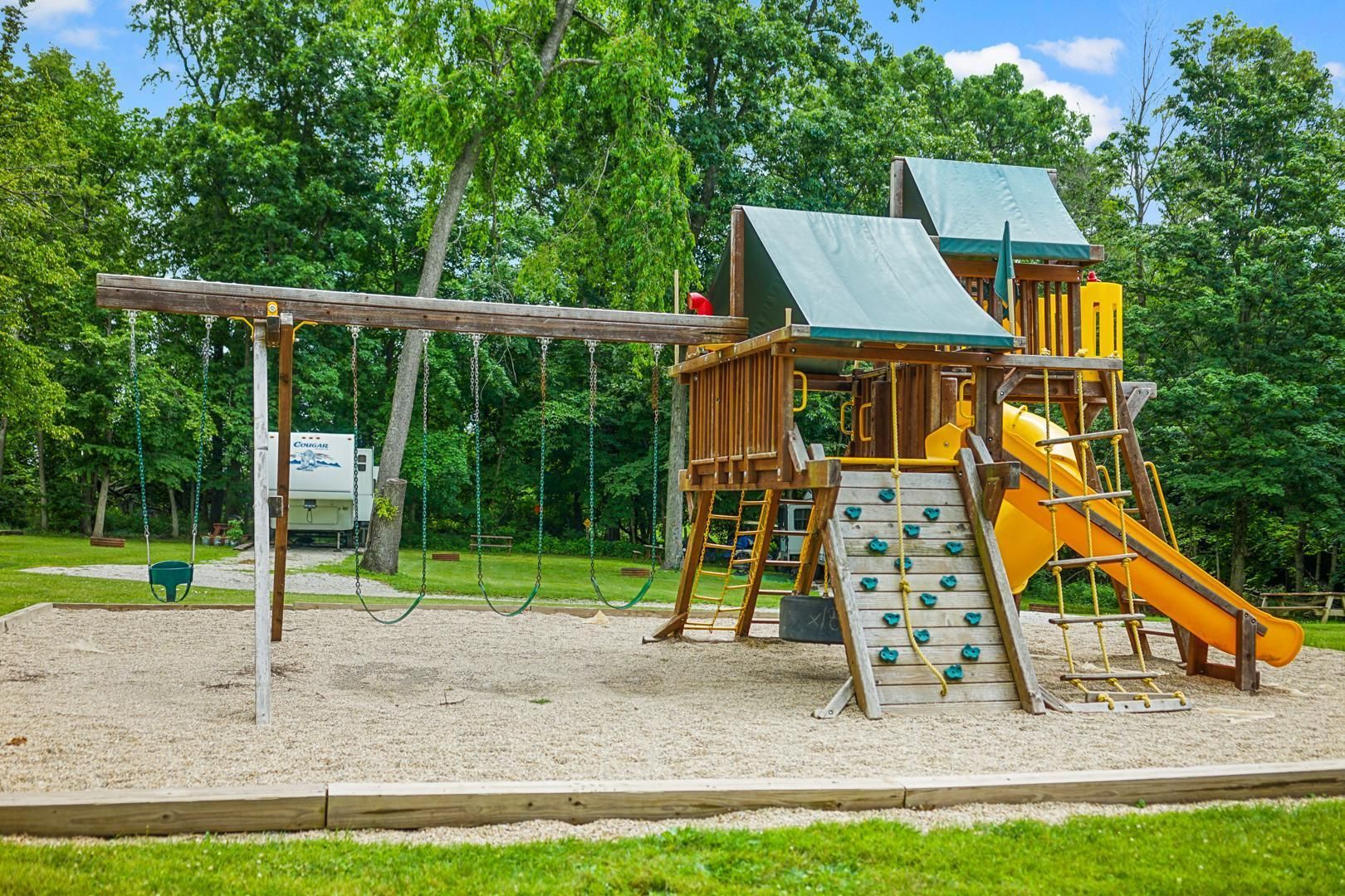 A wooden playground set with swings, a rock climbing wall, and a yellow slide on a wood-chip ground surrounded by trees.