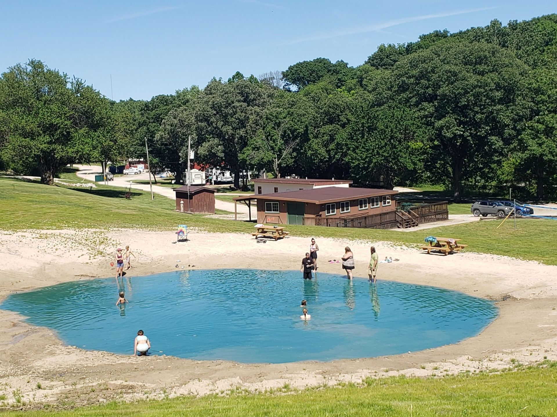 People wading in a small, bright blue circular pond surrounded by a sandy beach, green grass, and trees.