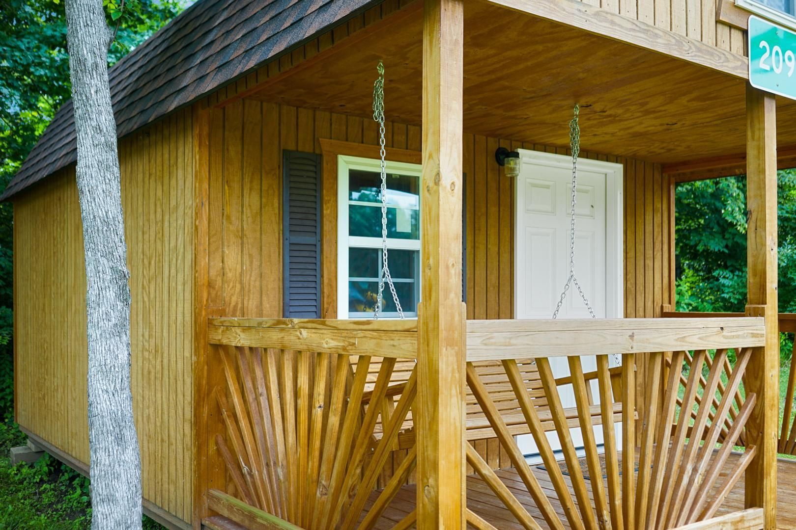 A rustic, wooden cabin with a porch featuring a sunburst railing and a white door, set in a wooded area.