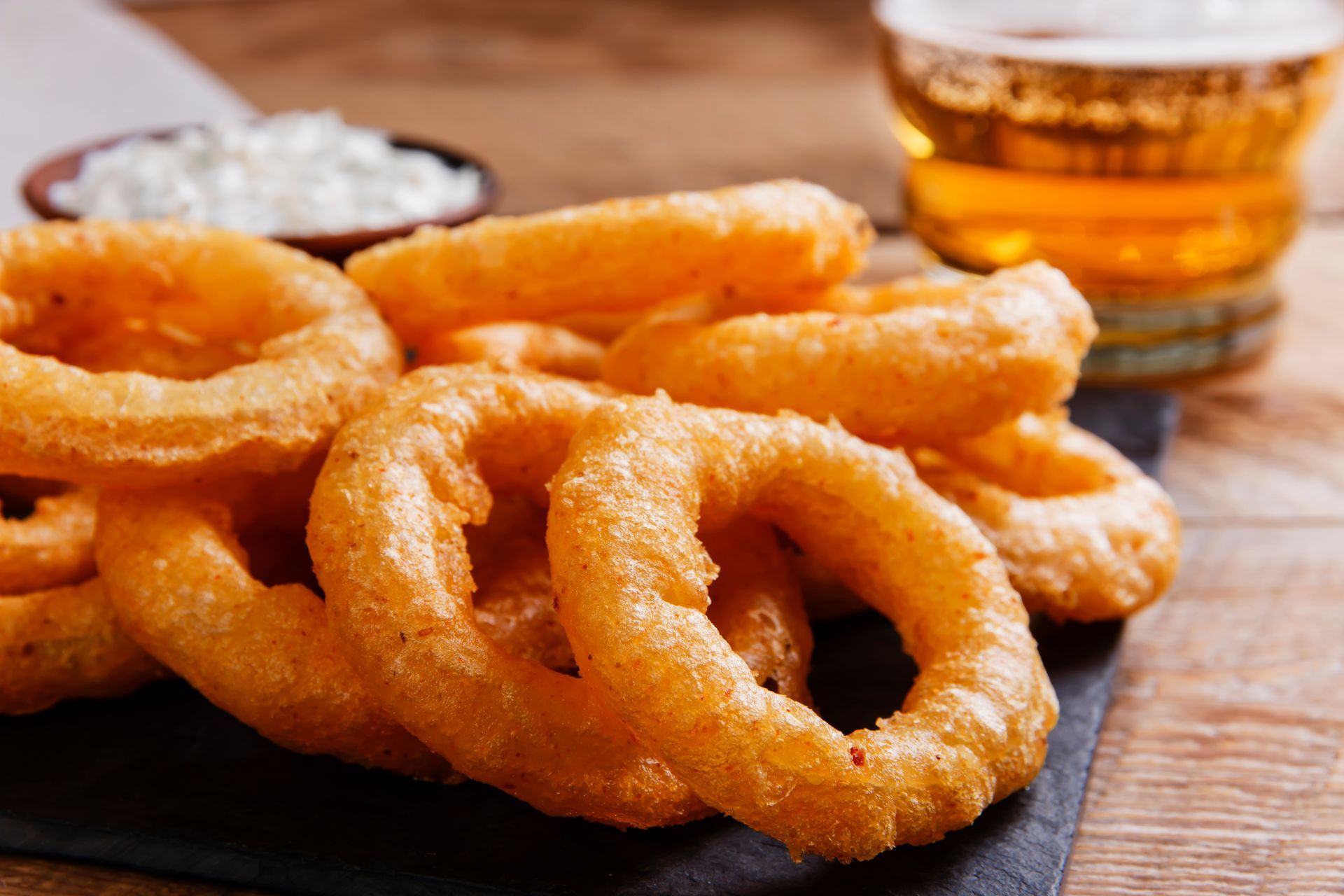 Golden-brown onion rings served on a dark slate board with a side of white dipping sauce and a glass of beer.