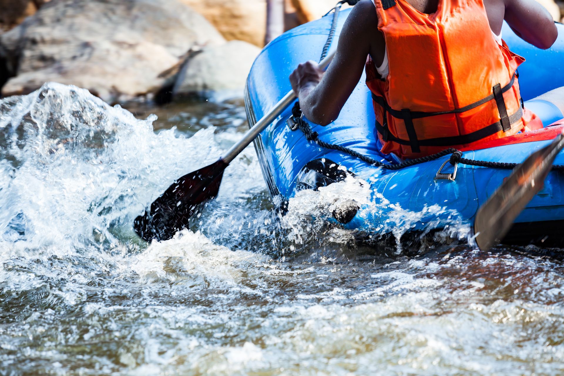 A person in an orange life vest paddles a blue raft through splashing river rapids.