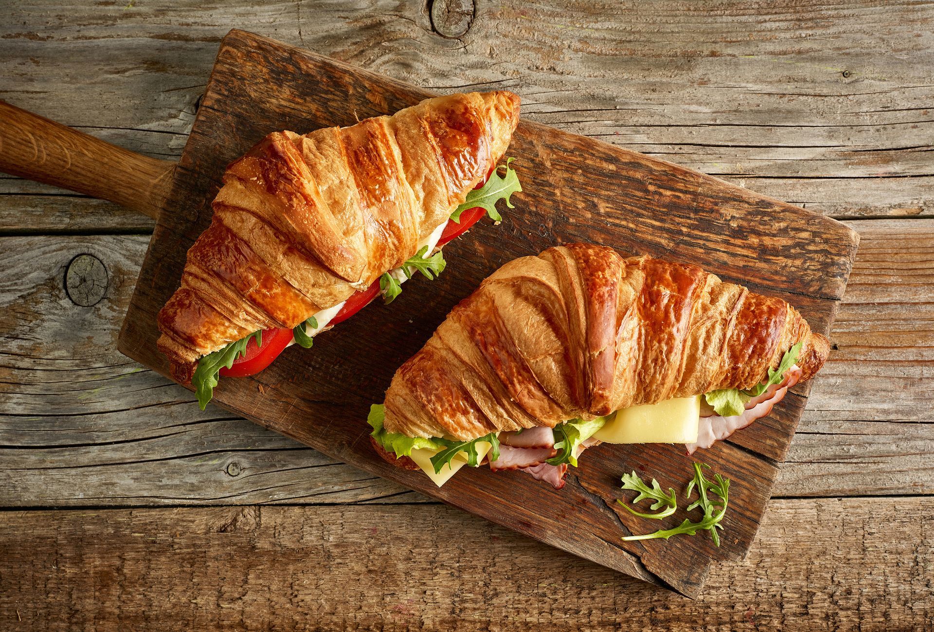 Two croissant sandwiches with ham, cheese, and lettuce on a rustic wooden cutting board.