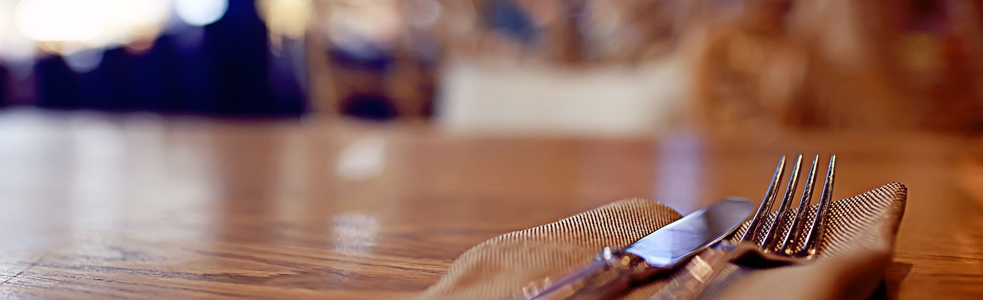 A silver fork and knife resting on a beige cloth napkin on a wooden table in a dimly lit restaurant.