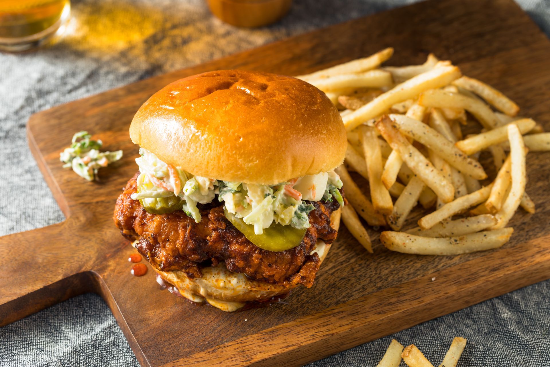 A fried chicken sandwich topped with coleslaw and a pickle, served with french fries on a wooden cutting board.
