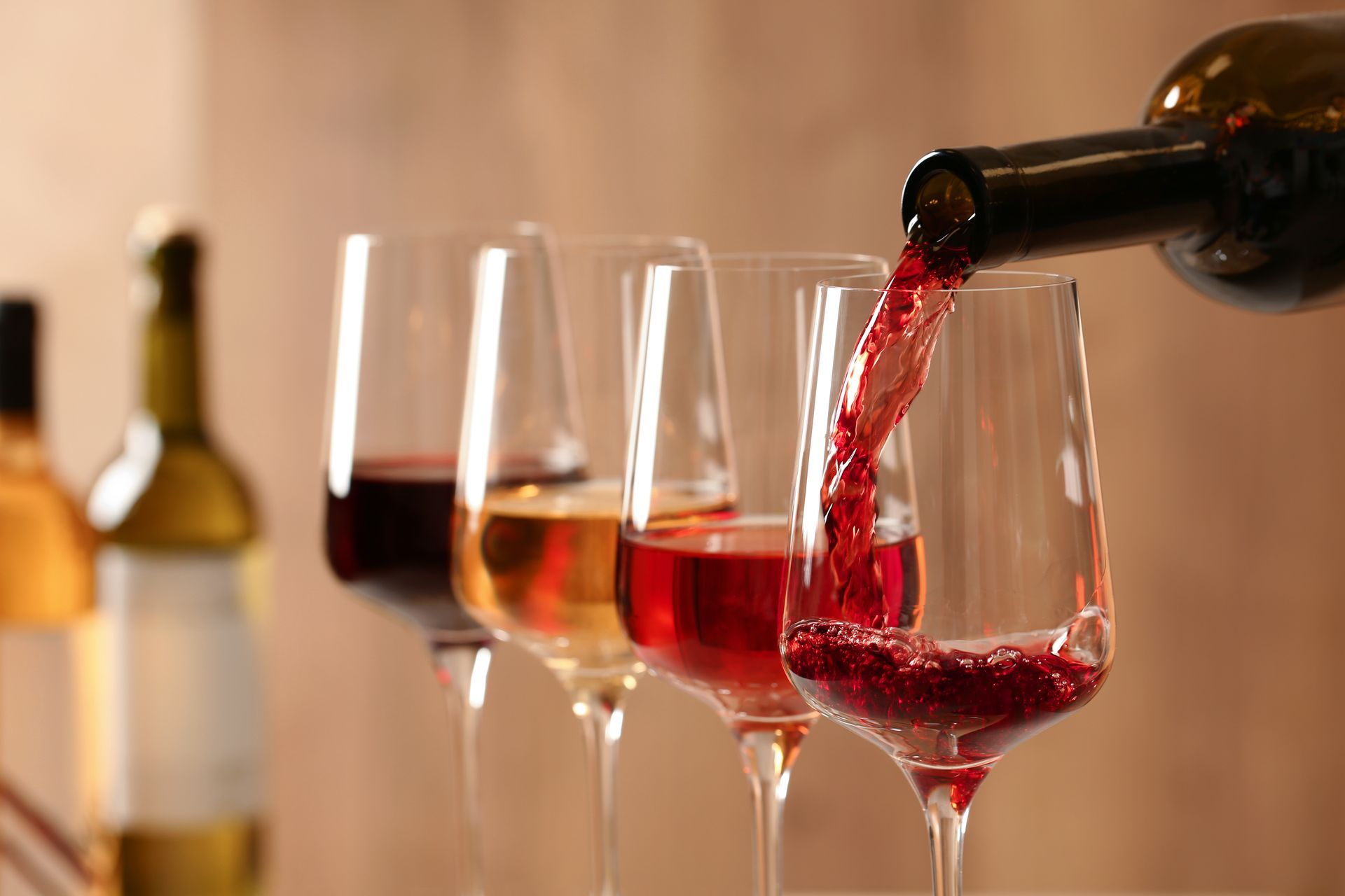Red wine pours from a bottle into a glass, with other filled wine glasses and bottles in the blurred background.