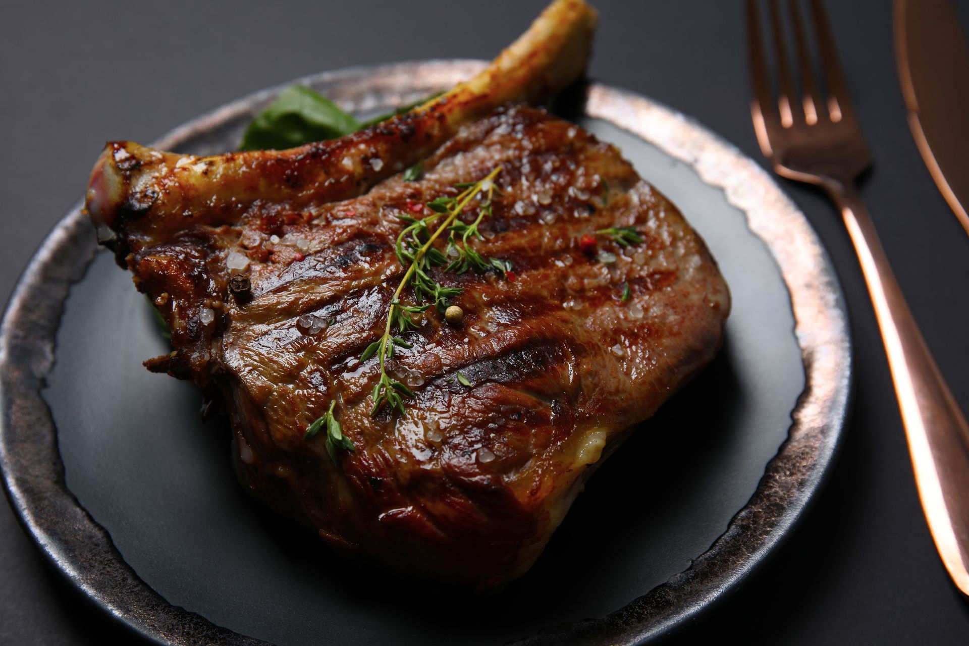 A grilled bone-in ribeye steak topped with fresh herbs, served on a dark plate next to a copper fork and knife.